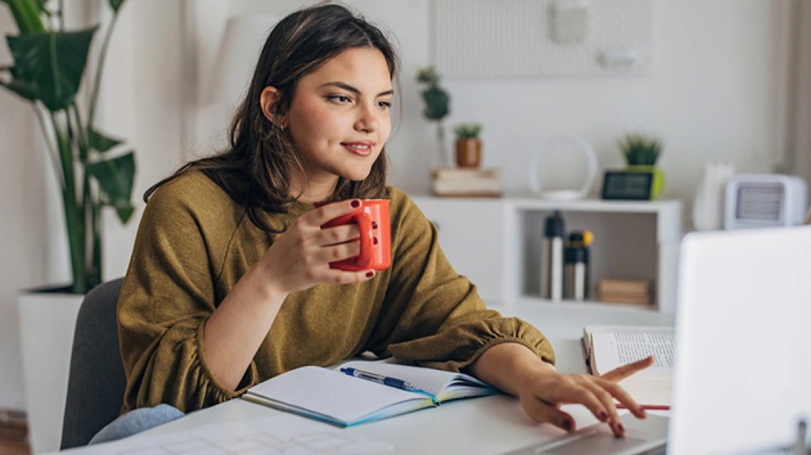 Student holding coffee mug and working on laptop in home setting